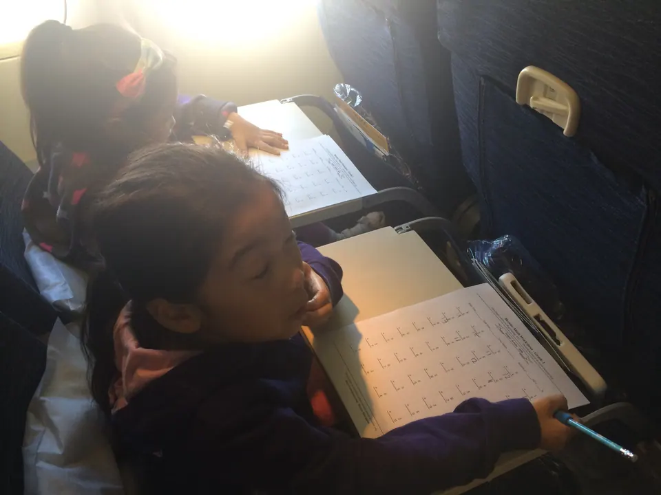Two girls in purple jackets bend over printed worksheets on their tray tables in an airplane seat row, sunlight coming in from the window.