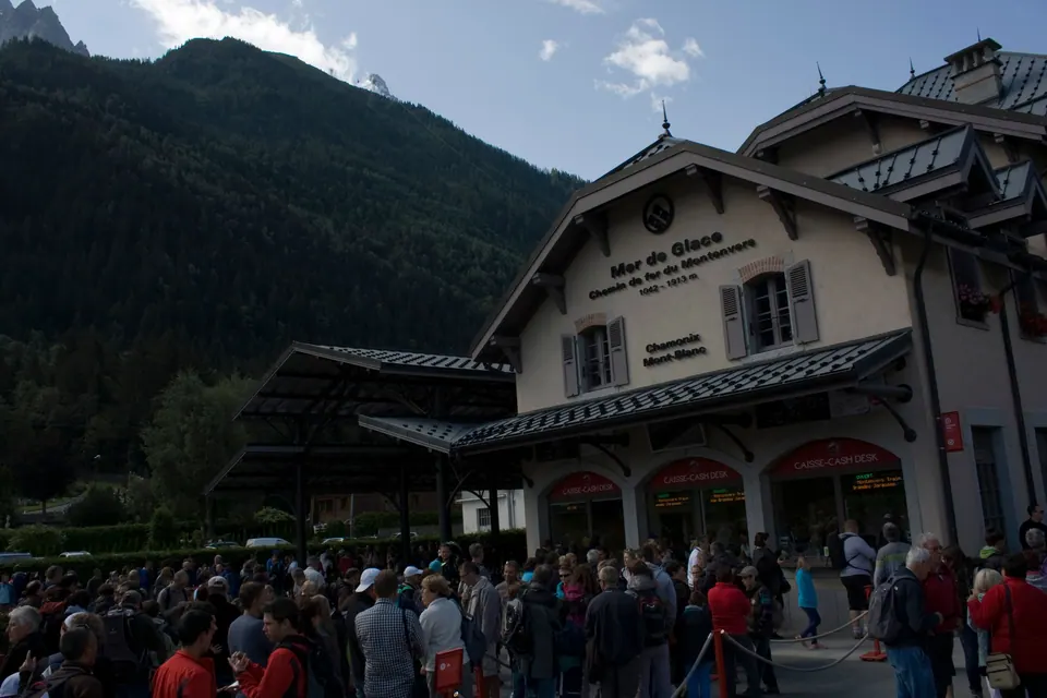 A large crowd waits outside a chalet-style station building whose sign reads Mer de Glace, with a dark forested mountainside behind it.