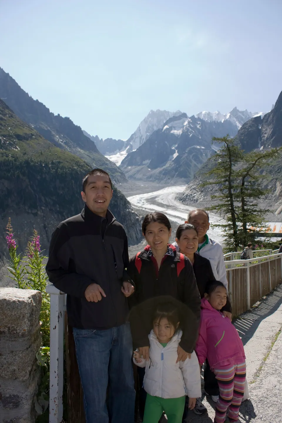 Four adults and two girls pose at a wooden railing in bright sun; behind them a gray glacier runs down a deep valley between jagged, snow-streaked peaks.