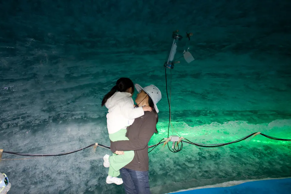 In a tunnel of rippled ice lit blue and green, an adult in a white cap holds a small girl up against the ice wall.