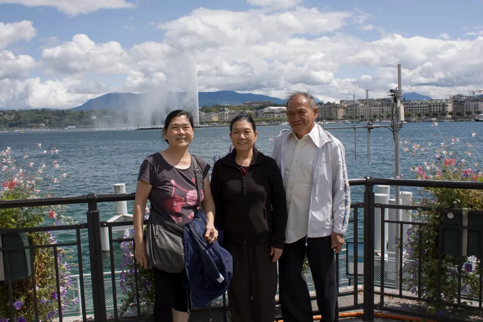 Three adults stand at a railing hung with flower boxes; behind them a tall fountain rises from the lake in front of the city skyline under puffy white clouds.