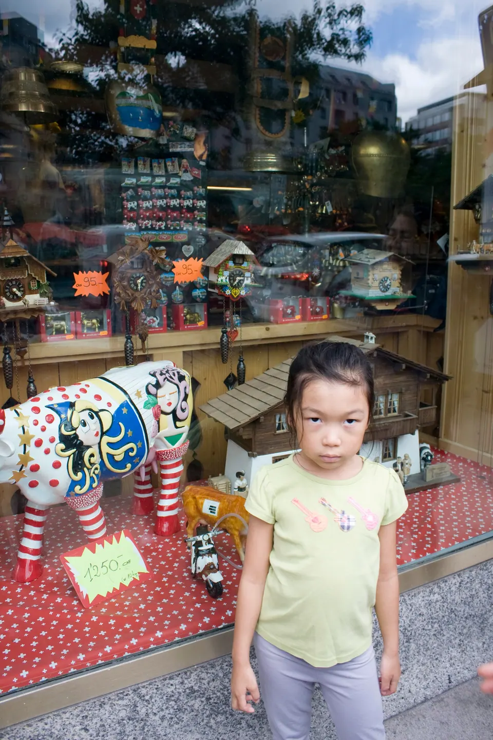 A girl in a lime T-shirt stands in front of a shop window full of wooden toys, cuckoo clocks, a toy chalet and a painted cow figure.