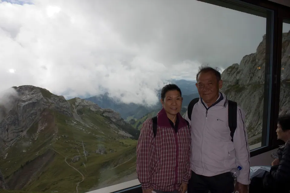 Two older adults stand at a wide window; behind them green ridges and rocky peaks disappear into white cloud.