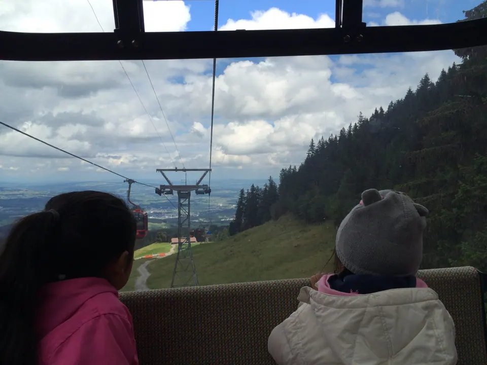 Two girls, one in a knit hat, look out of a cable car window; the cable drops past a pylon and a red cabin over green pasture and forest toward a wide plain.
