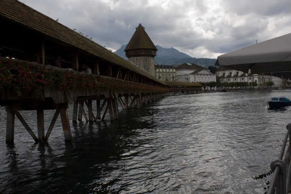 A long covered wooden bridge hung with flower boxes crosses a river to an octagonal stone tower under a dark, cloudy sky.