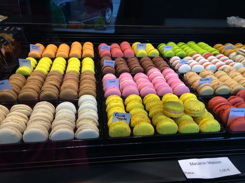 A display case holds neat rows of macarons in orange, yellow, pink, brown, green and white, with small flavor labels and a price card reading Macaron Maison, 1.00 euro.