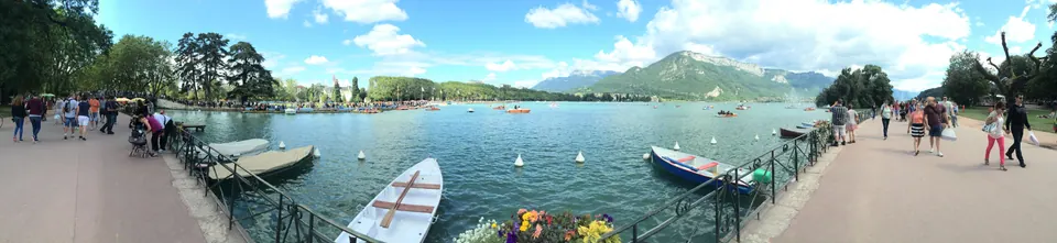 A wide panorama of a clear green lake with moored rowboats below a stone quay, people strolling along both sides, and mountains under a blue sky with white clouds.