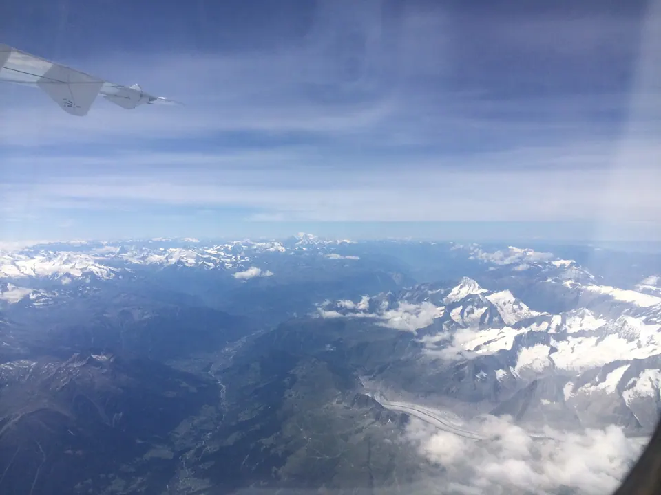 Snow-covered Alpine peaks, ridges and a long glacier spread out below a plane window under a pale blue sky, with the tip of the wing at the top left.