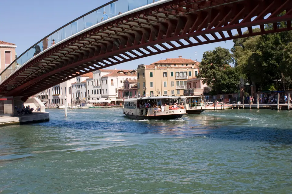 A crowded water bus passes under the long, low arch of a modern steel-and-glass bridge over a green canal, with pastel palazzi in the sun behind.