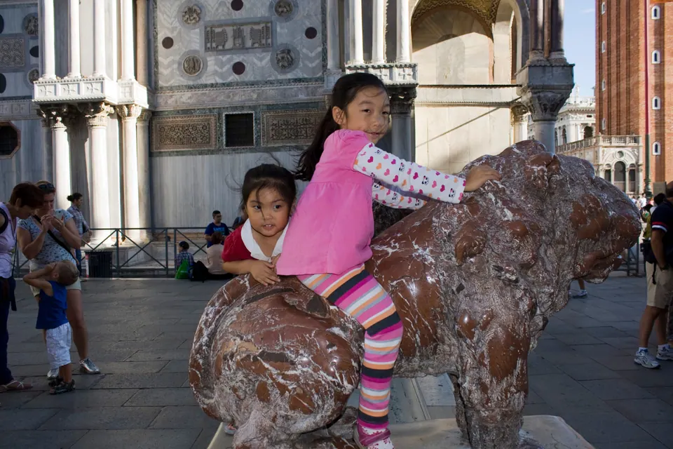 Two girls sit astride a worn red marble lion statue on a stone square, the carved marble corner of a basilica and a brick bell tower behind them.