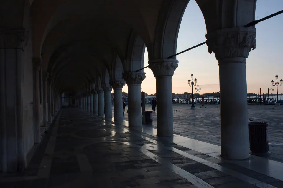 A long colonnade of white stone columns and pointed arches in shadow, framing lamp posts on the waterfront and a pale pink dawn sky.