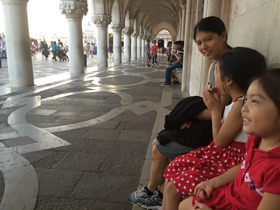 A woman and two smiling girls in red dresses sit against a pillar on a patterned stone floor, the columns of the arcade and a crowd running off into the distance.