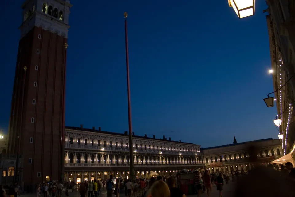 A tall brick bell tower and long lit arcades frame a crowded square under a deep navy sky.