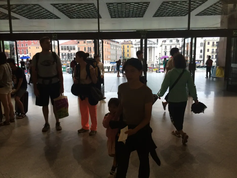 People with bags walk in silhouette through a station hall with a glass front; the buildings along a canal glow in daylight outside.