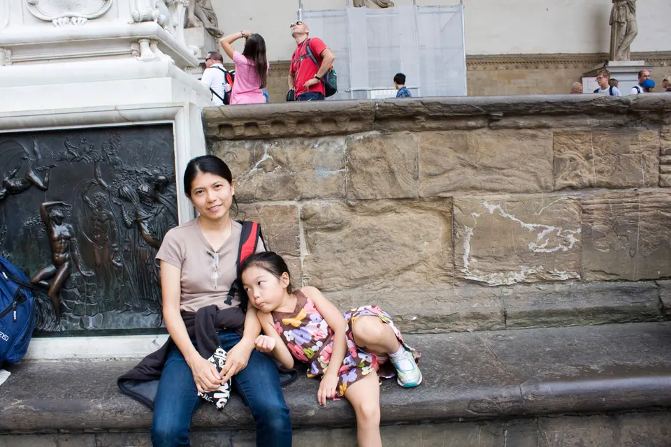 A woman and a girl in a flowered dress sit on a stone ledge beside a bronze relief at the foot of a statue's plinth, tourists standing above them.