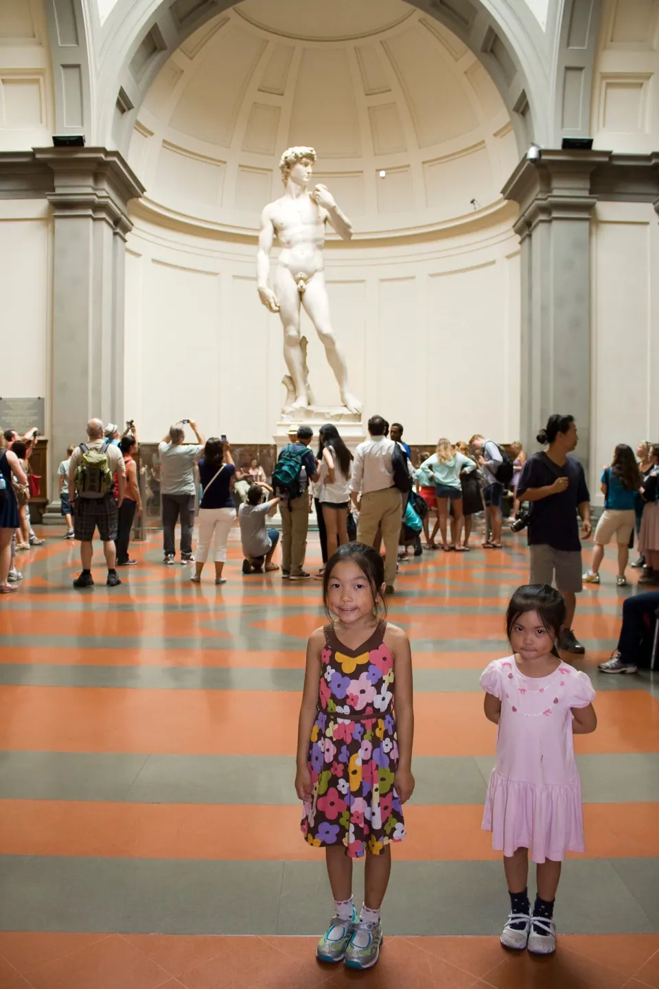 Two girls in summer dresses stand on a striped floor; far behind them a white marble statue of a standing nude figure rises under a domed, skylit alcove, surrounded by visitors.