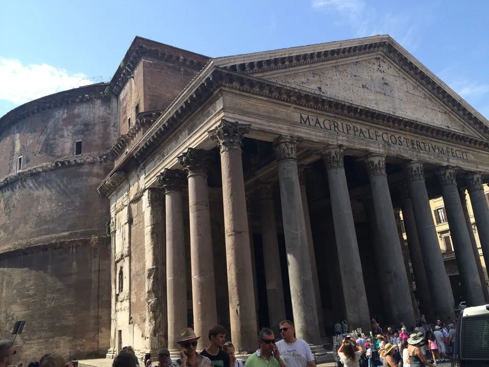 A massive temple portico of tall granite columns under a triangular pediment inscribed M AGRIPPA, against a blue sky, with a crowd of tourists at its foot.
