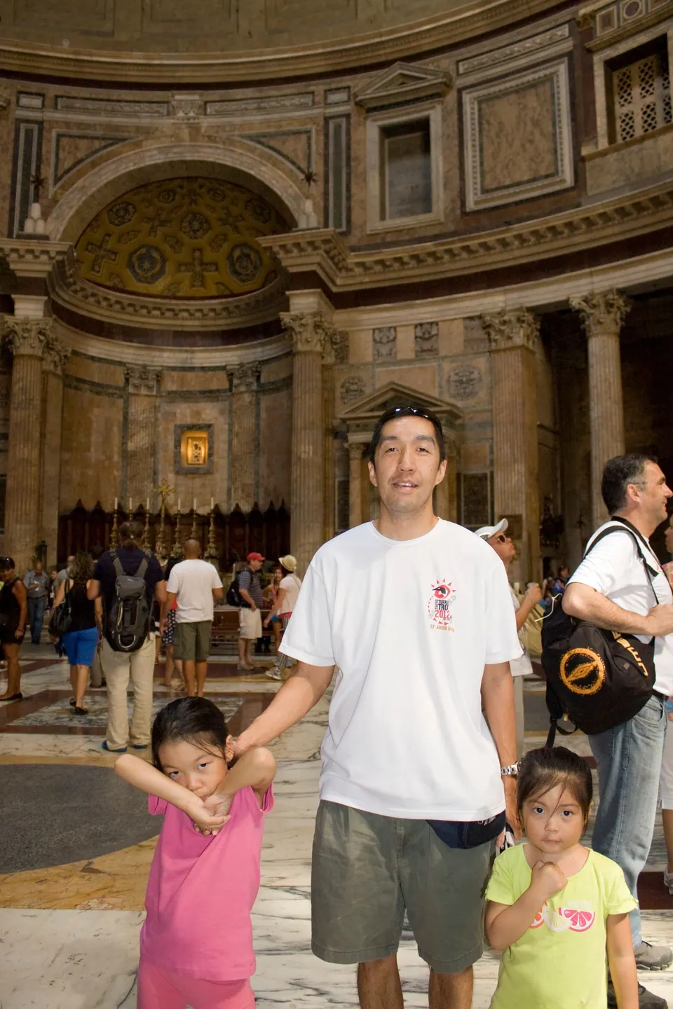 A man in a white T-shirt holds hands with two small girls on a marble floor, the curved coffered walls and a gilded apse of a domed interior behind them.
