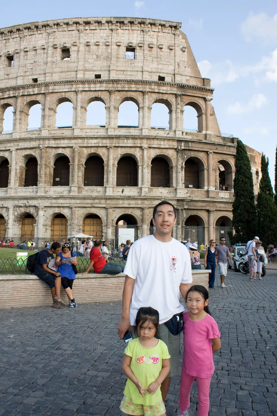 A man and two girls stand on cobblestones in front of the tiered arches of the Colosseum in warm evening light.