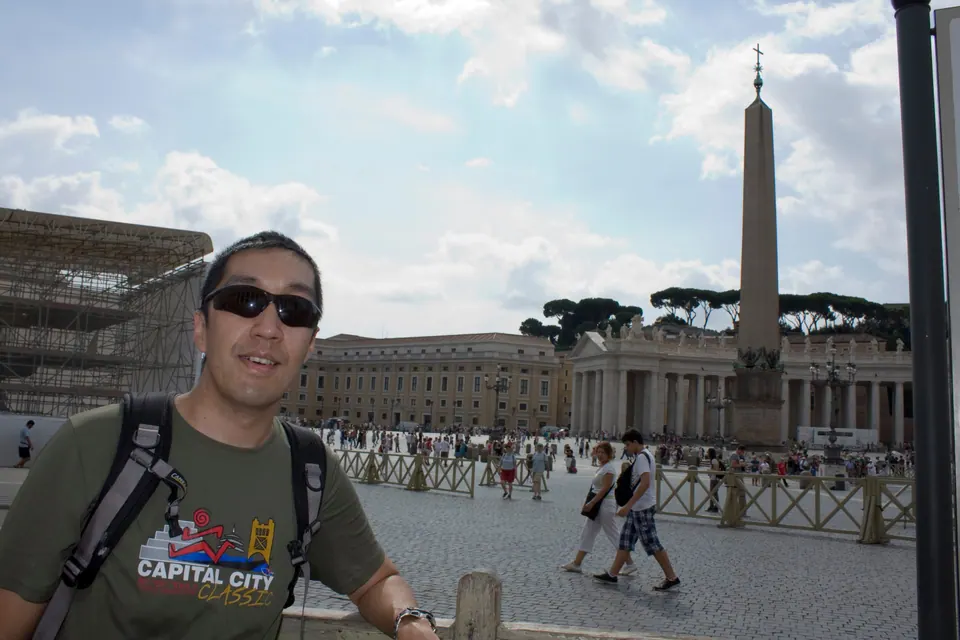 A man in sunglasses and a backpack smiles at the camera on a wide paved square, with an obelisk and a curved colonnade behind him under a cloudy sky.