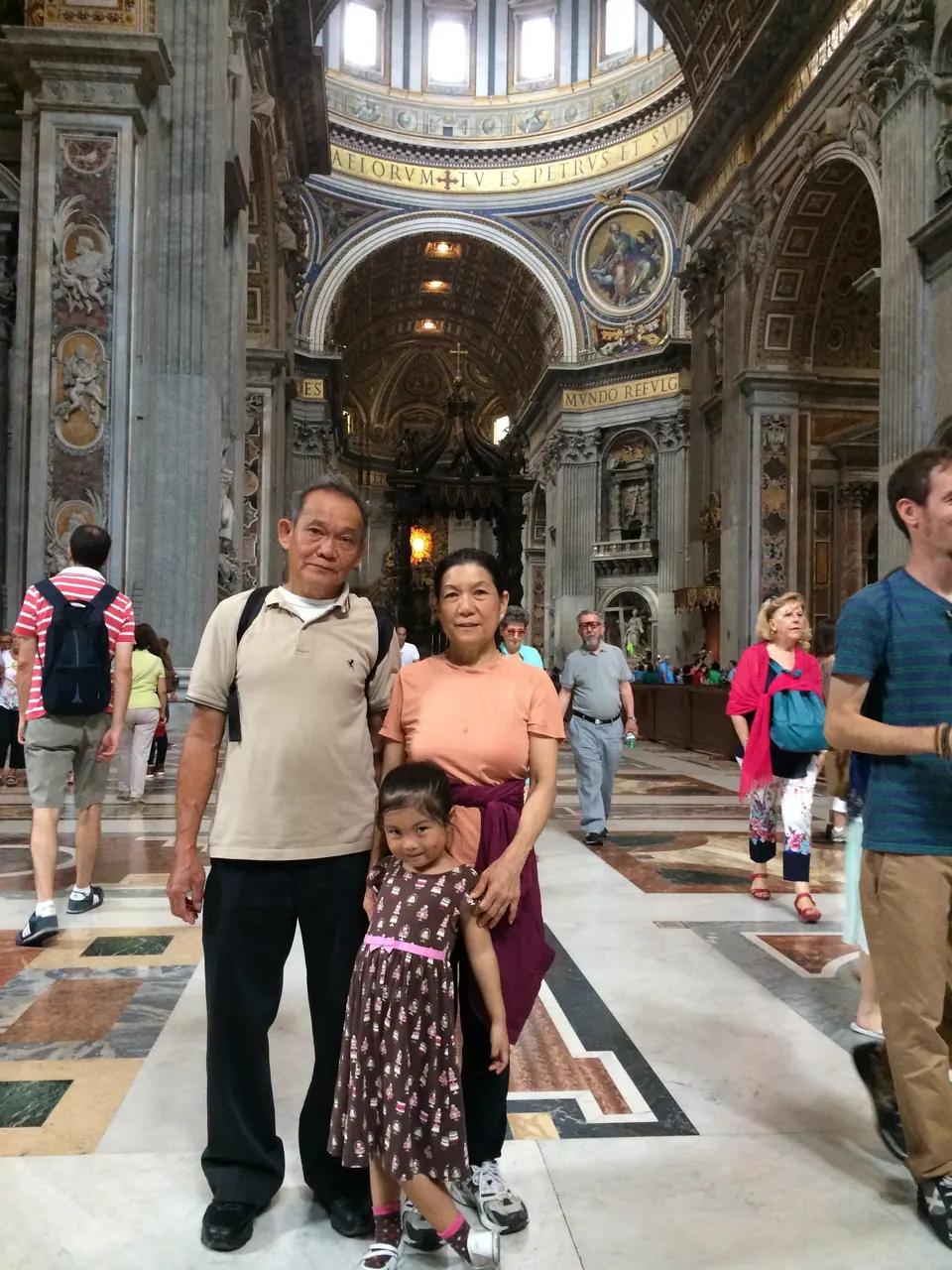 An older man and woman stand with a small girl on a colored marble floor in a vast church nave, gilded arches and the canopy over the altar behind them.