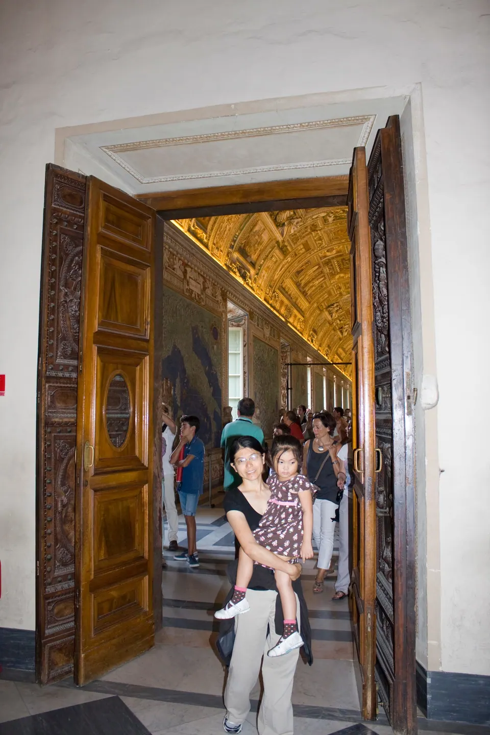 A woman holding a small girl stands in a tall carved wooden doorway that opens onto a long gallery with a gilded, painted ceiling, crowded with visitors.