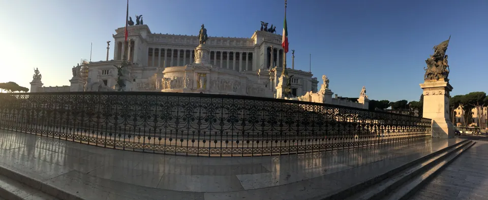 A wide panorama of a huge white marble monument with columns and bronze statues, lit by low sun, behind an iron railing and an empty paved square.