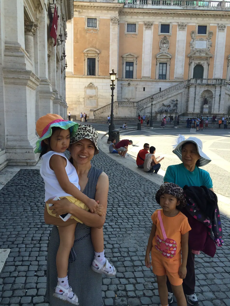 Two women in sun hats, one holding a small girl, stand on a cobbled square with another girl in orange; a palace with a double staircase is behind them.