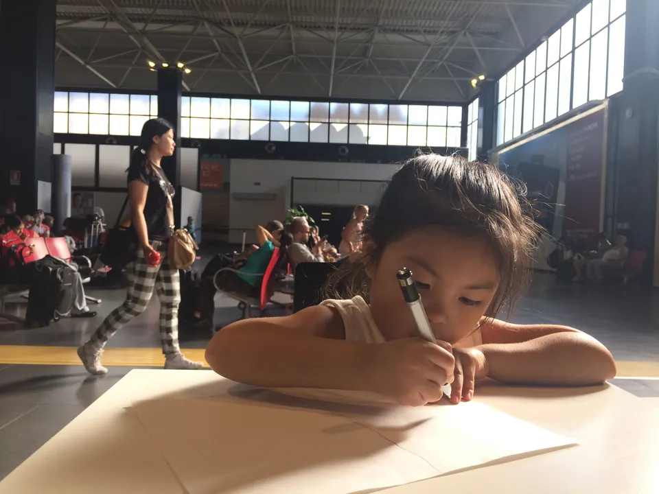 A girl rests her chin on her arms, drawing with a pen on a sheet of paper at a low table in a sunlit departure hall while a woman walks past.