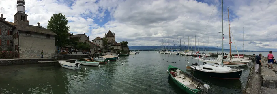 A wide panorama of a small harbor: medieval stone houses and a church tower on the left, rowboats and sailboats on green water, and big clouds over the lake.