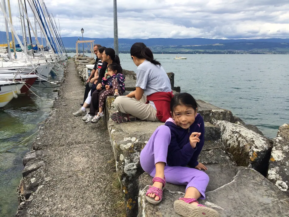 Two girls and a woman sit on the rough stone blocks of a jetty beside moored sailboats, with gray-blue water and a far shore behind them.
