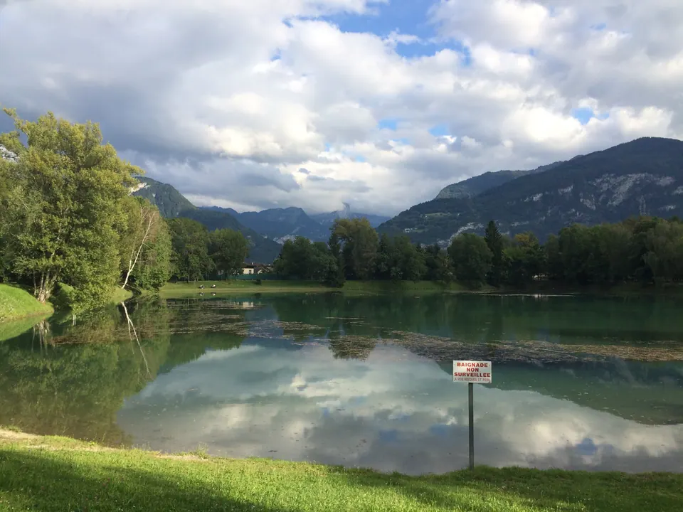 A small lake reflects white clouds, trees and wooded mountains; a sign on a post stands at the water's edge.
