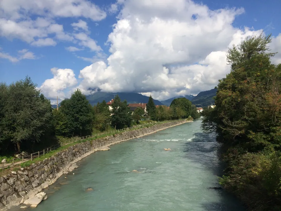 A milky green river runs between a stone embankment and trees toward mountains under big white clouds.