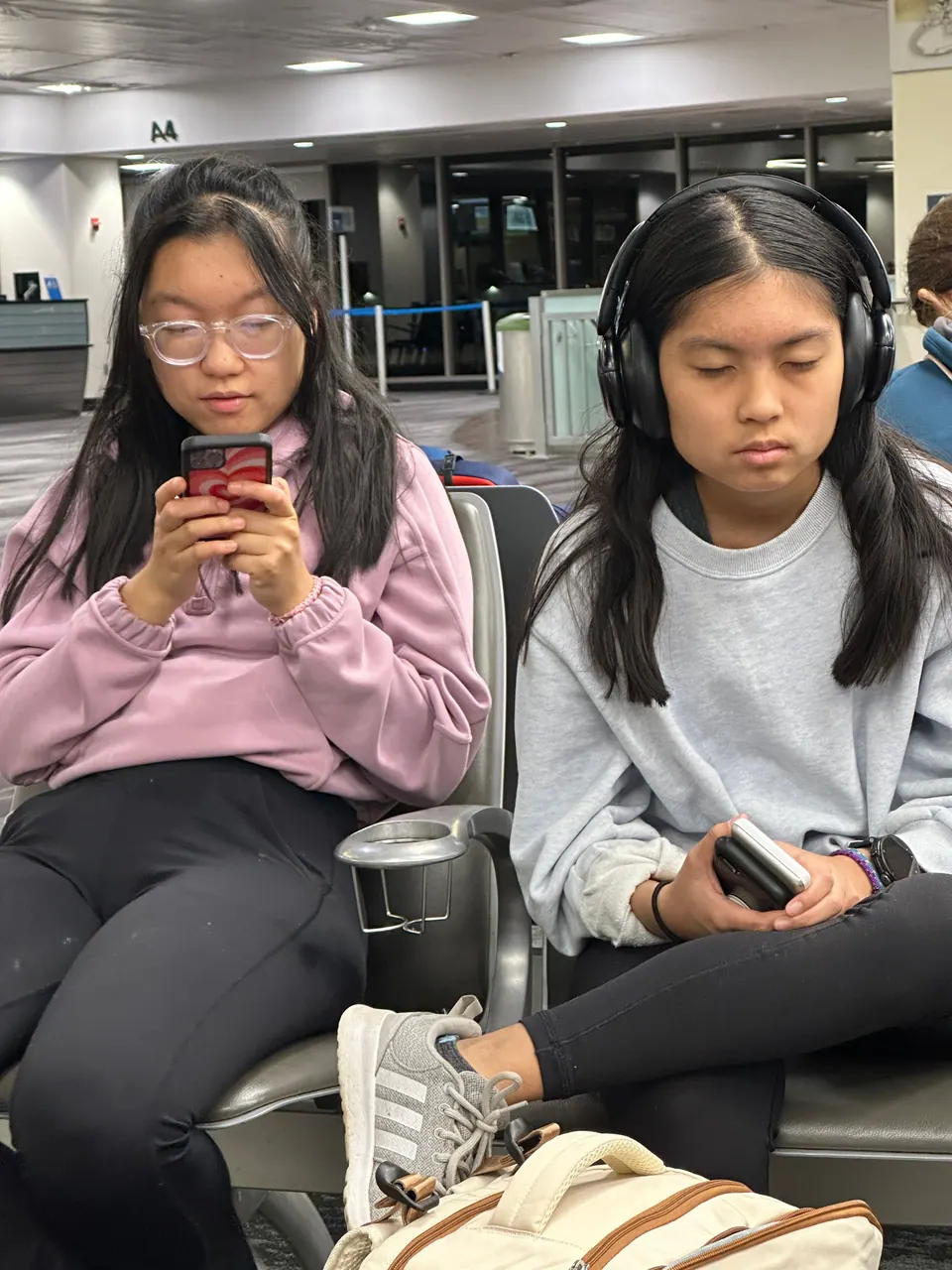 Two girls sit side by side on gate-area seats at night: one in a lilac hoodie and glasses looks at her phone, the other in a gray sweatshirt and over-ear headphones holds a phone in her lap, a tan backpack in front of them.