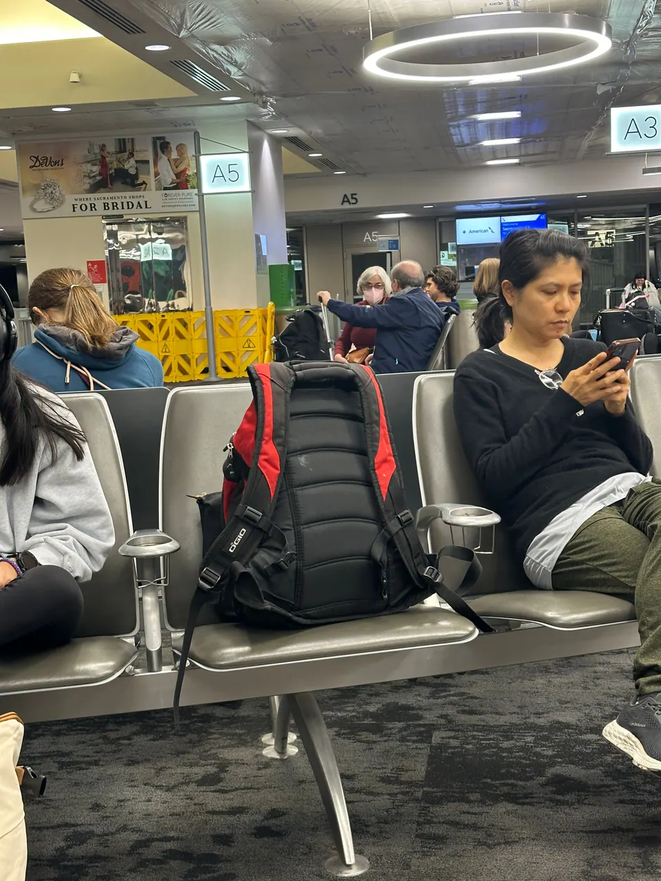 A woman in a black top looks at her phone in an airport gate area, a red and black backpack on the seat beside her.