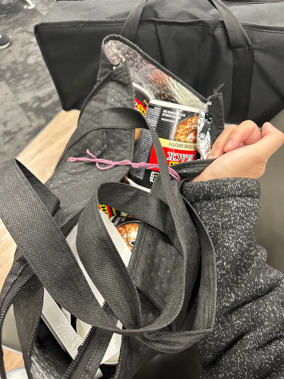 A hand holds open a black tote bag at the airport; inside are instant-noodle cups and snack packets.