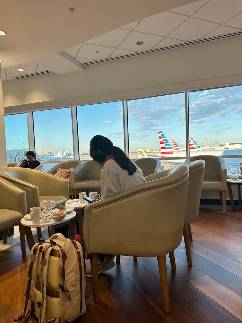 A girl sits in a tan armchair by floor-to-ceiling windows looking out at the airfield, where several American Airlines tails show; a coffee cup and a backpack are beside her.