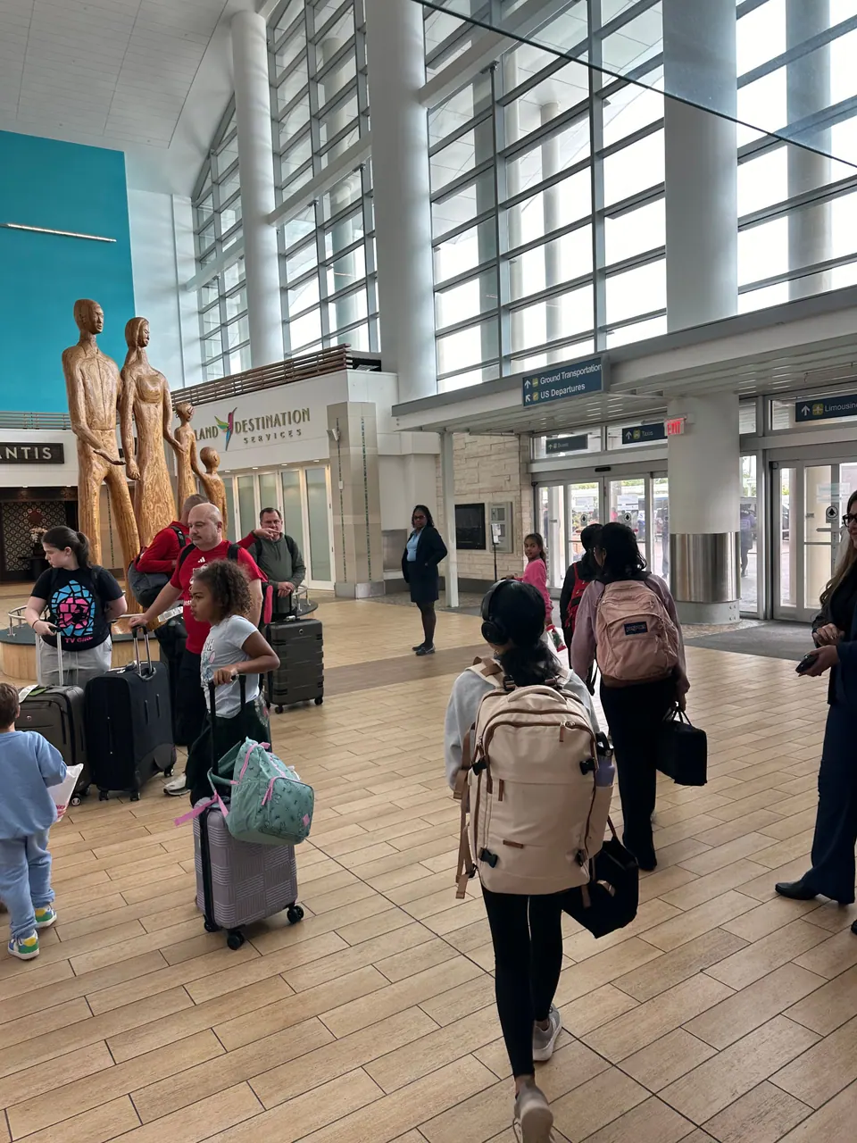 Travellers with wheeled suitcases and backpacks cross the tiled arrivals hall of an airport, past a group of tall carved wooden figures and a turquoise wall.