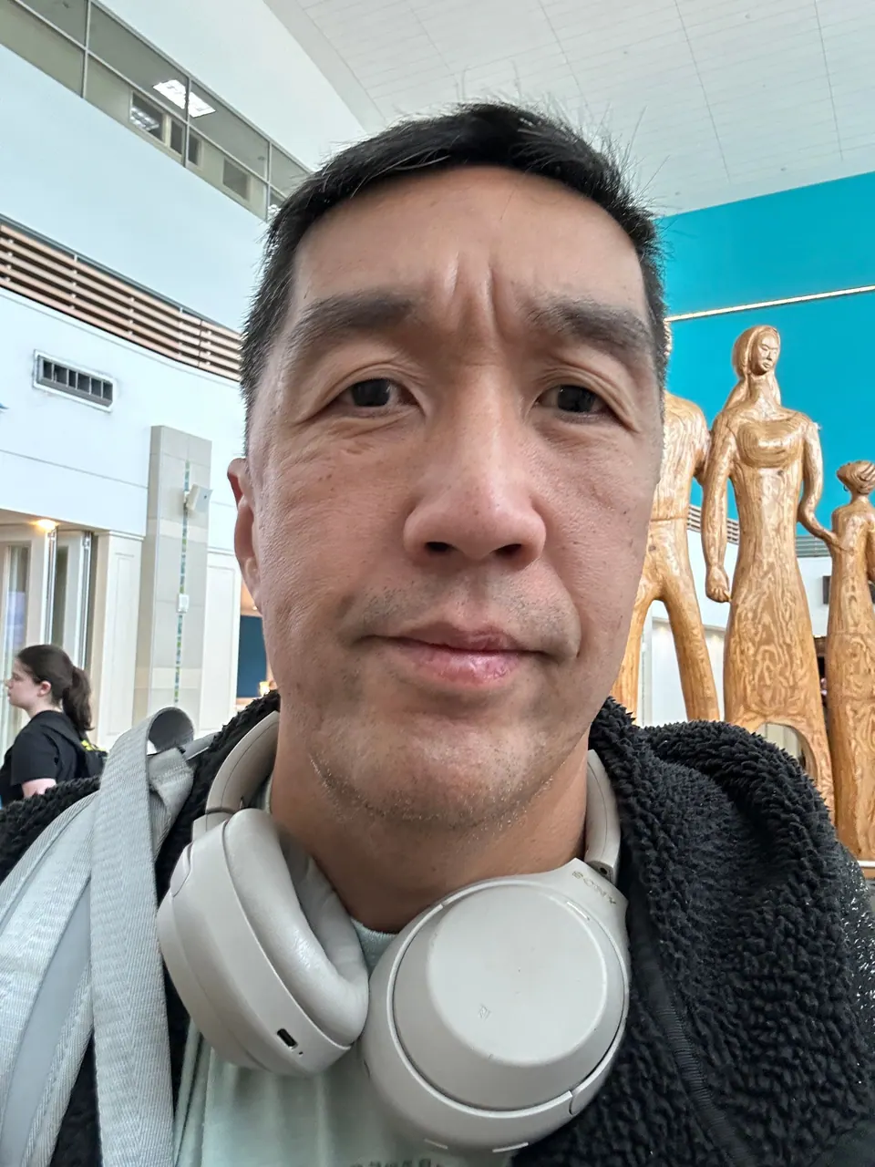 A man with headphones around his neck takes a selfie in a bright terminal hall, tall golden carved figures behind him.