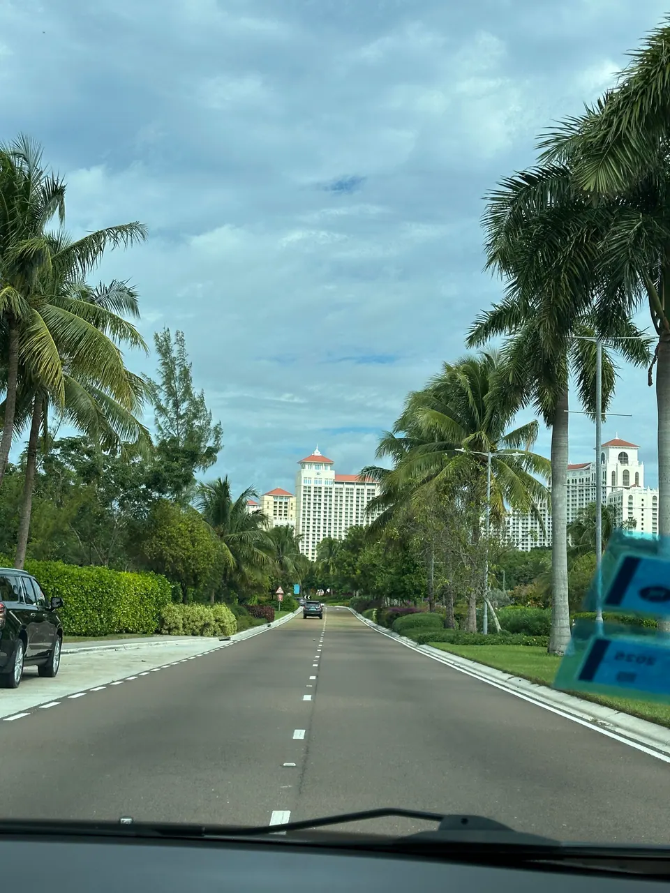 Through a taxi windshield, a straight road lined with tall palms and clipped hedges runs toward the white towers of a resort in the distance.