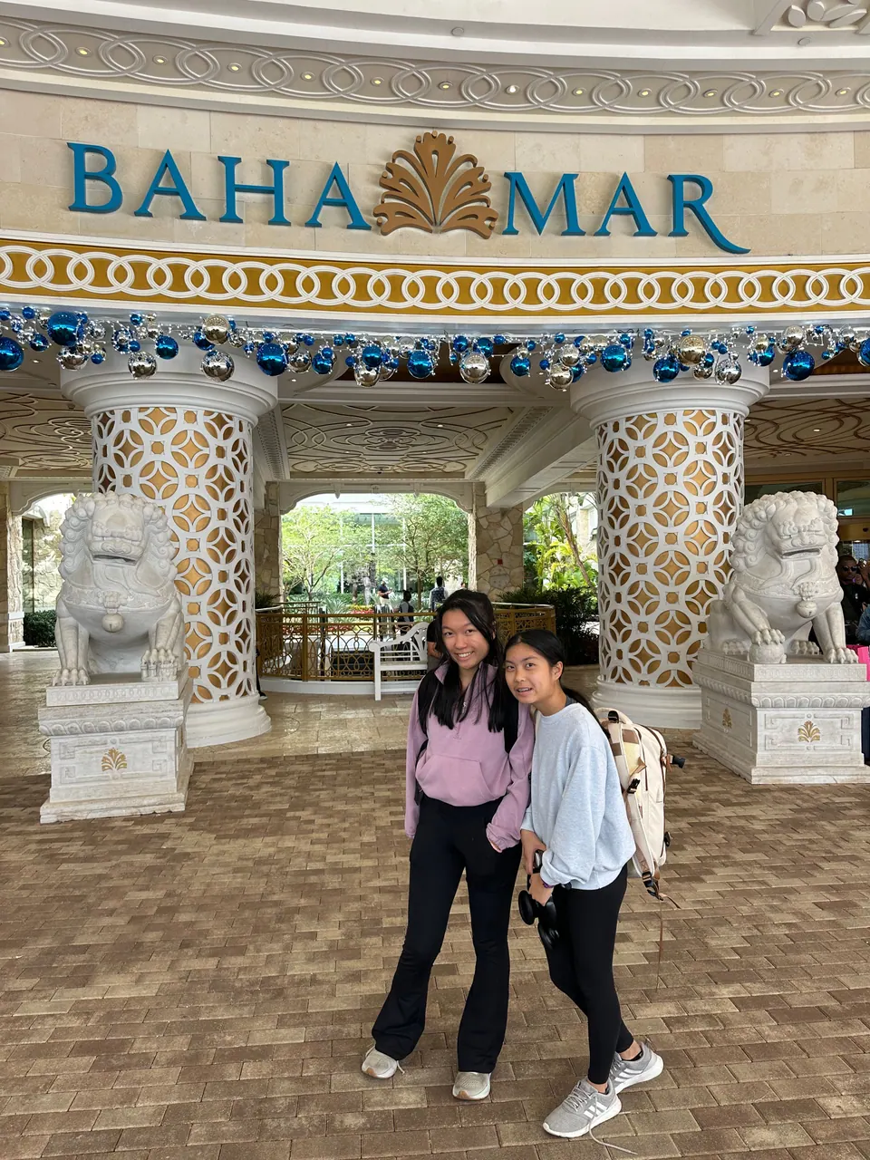 Two girls pose arm in arm on a brick driveway beneath the BAHA MAR sign, between white lattice columns and two stone guardian-lion statues, a blue and silver holiday garland overhead.