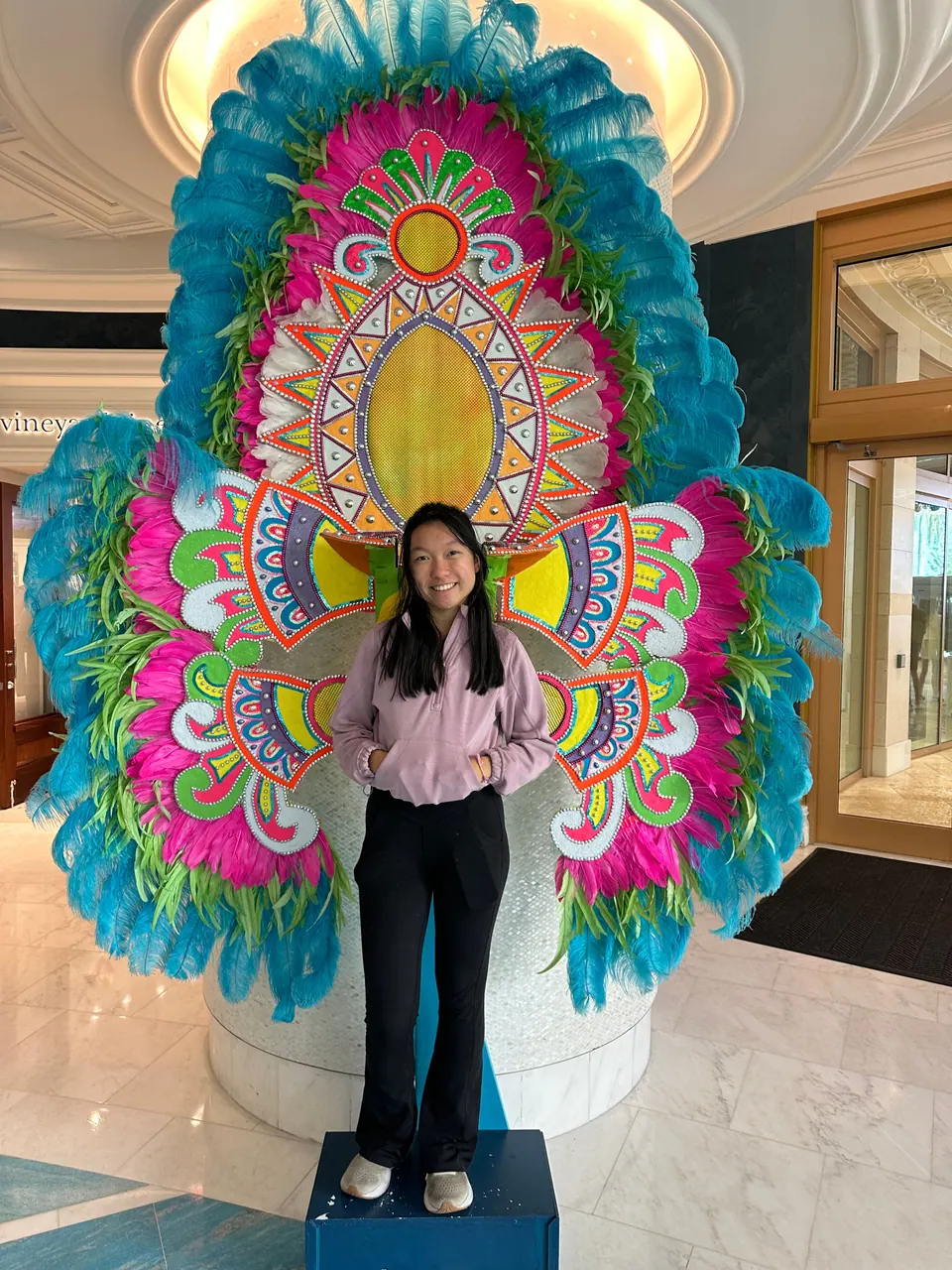 A girl in a lilac hoodie and black trousers stands on a low blue platform in front of a huge feathered carnival-style costume in bright pink, yellow and teal, in a marble hall.
