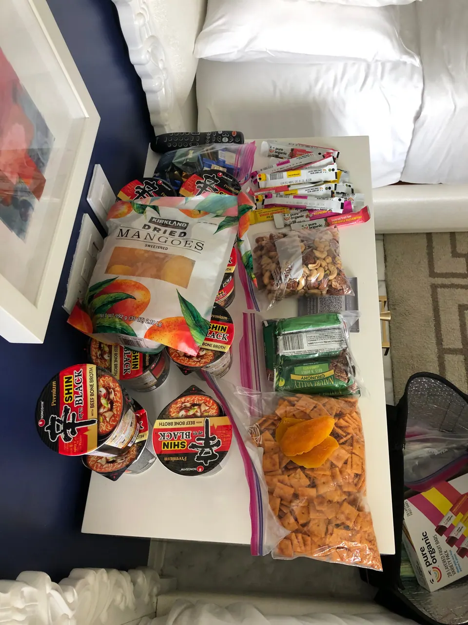 Bags of dried mangoes and other snacks, several instant-noodle cups and a zip bag of chips are spread out on a white surface in a hotel room.