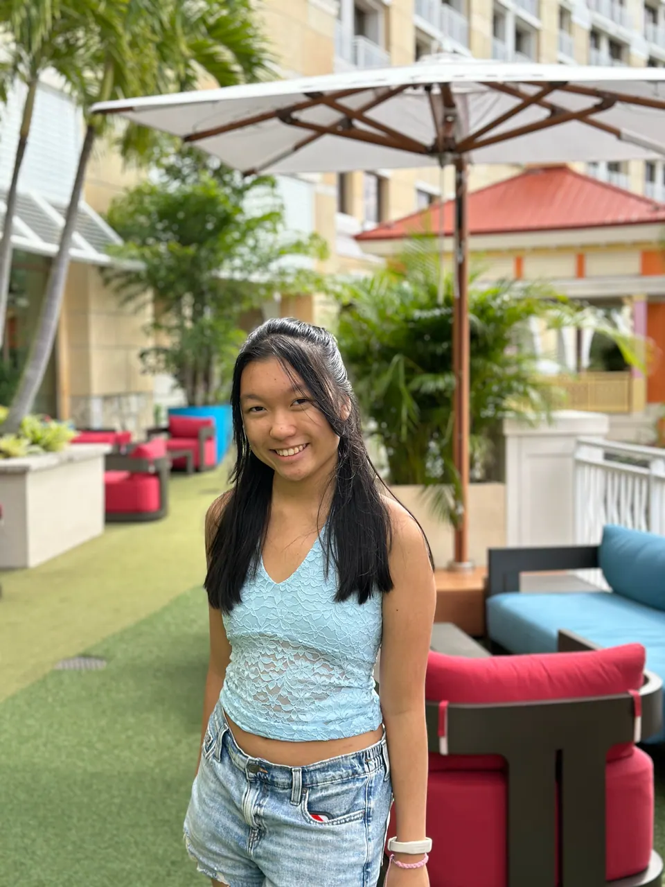 A smiling girl in a light-blue lace top stands on a lawn beside red and blue lounge chairs and a market umbrella, resort buildings and palms behind her.