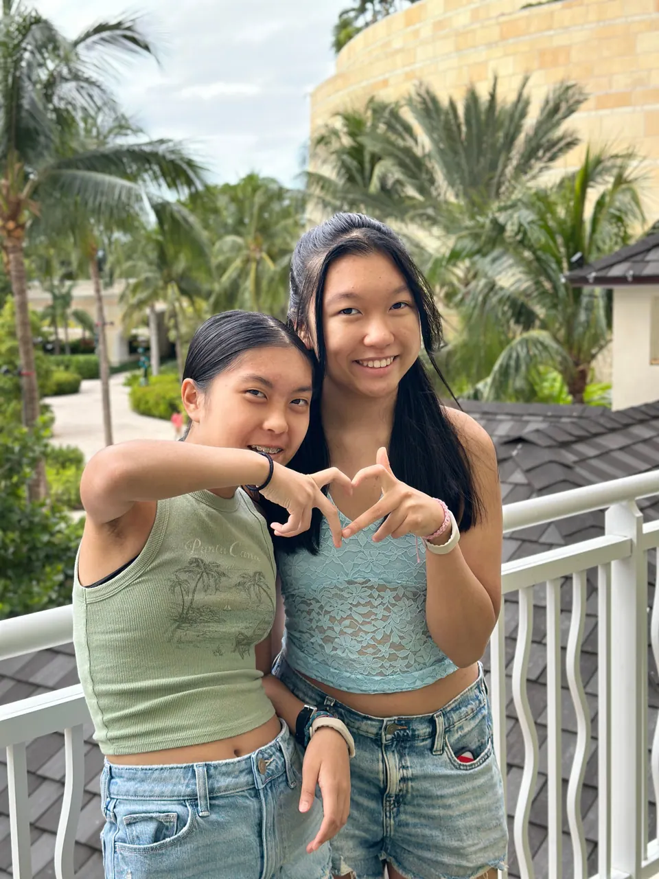 Two girls lean together at a white balcony railing making a heart with their hands, palms and a landscaped garden behind them.