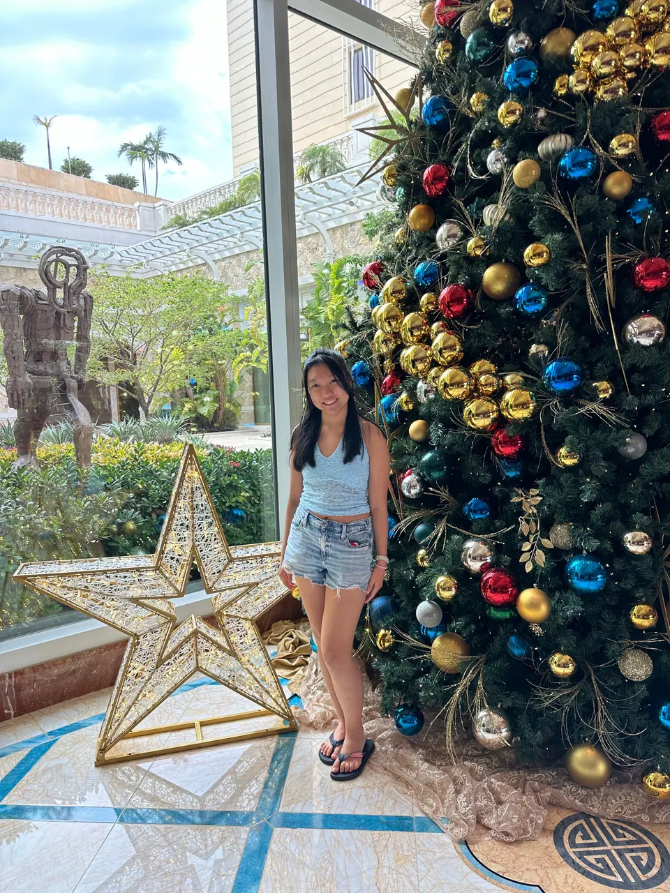 A girl in a light-blue top and denim shorts stands beside a tall Christmas tree hung with red, blue and gold ornaments and a large gold lattice star, beside a window onto a garden courtyard.