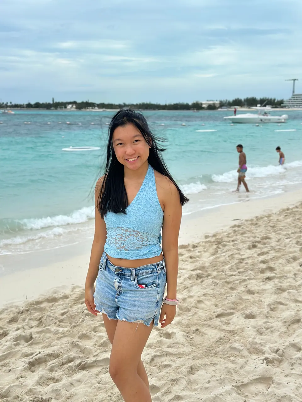A girl in a light-blue halter top and denim shorts stands on white sand at the water's edge, smiling, turquoise surf, a boat and swimmers behind her under a cloudy sky.