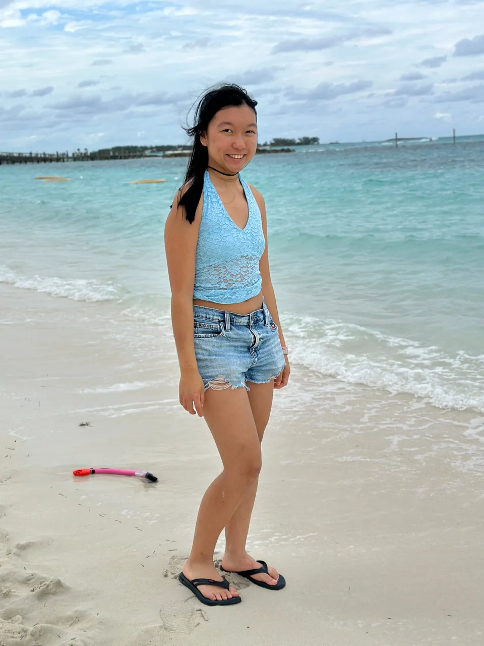 A girl in a light blue top and denim shorts stands on white sand at the water's edge, turquoise water and a low shoreline behind her under a cloudy sky.