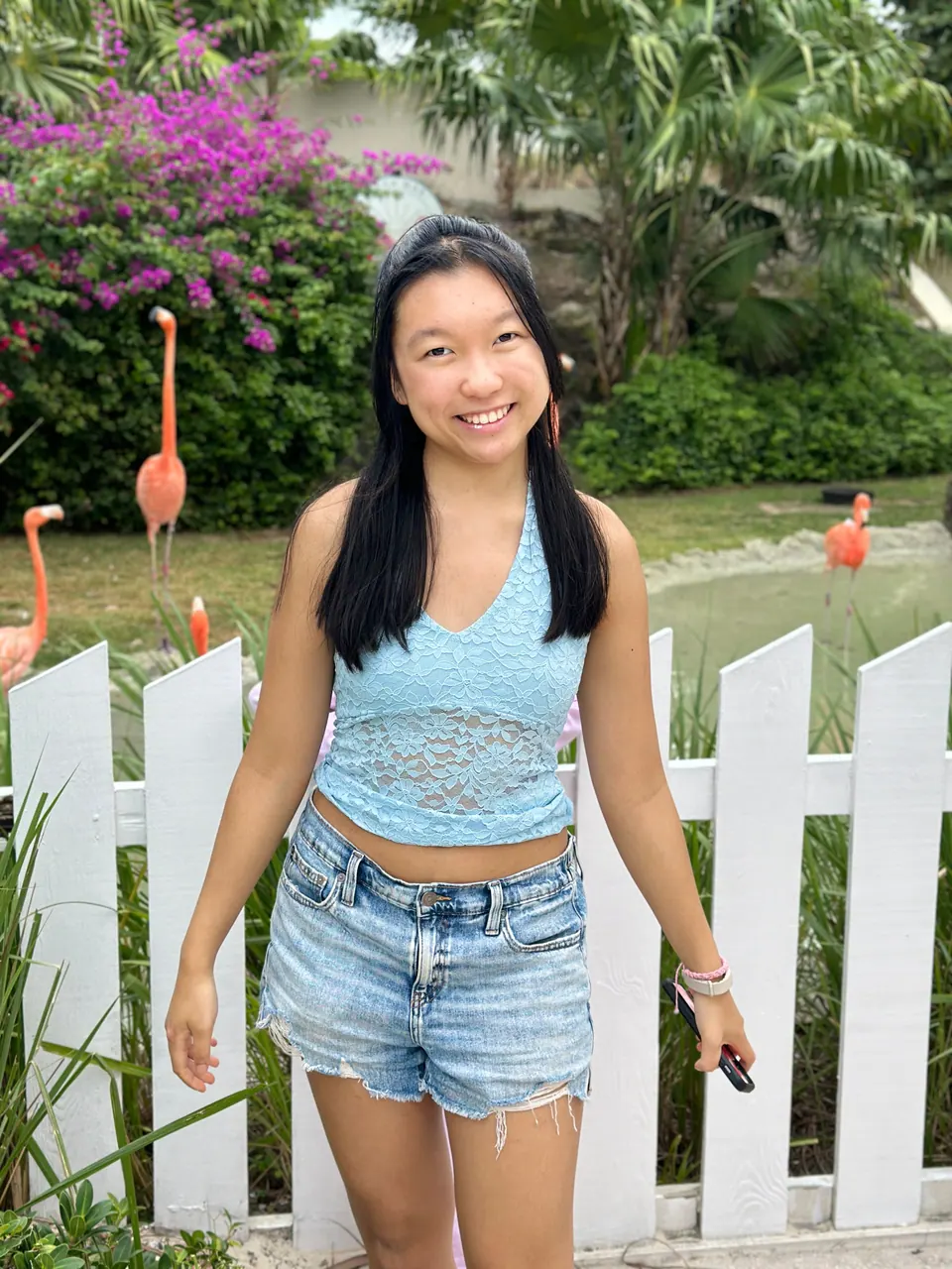 A girl in a light-blue halter top smiles in front of a white picket fence; pink flamingos stand on the grass behind it, under a bush of magenta bougainvillea.