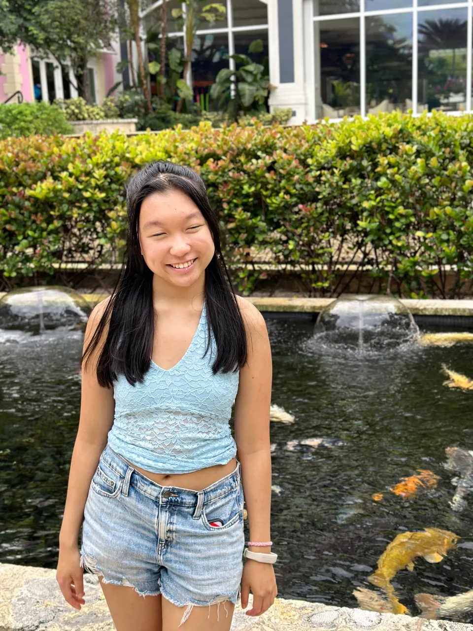 A girl stands beside a dark pond with orange and yellow koi and two small fountains, in front of a clipped hedge.